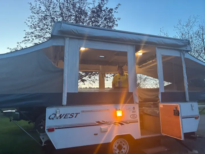 A person stands inside a lit Q-WEST pop-up camper, an inviting RV rental for evening adventures under a twilight sky.. Jayco Qwest 2004