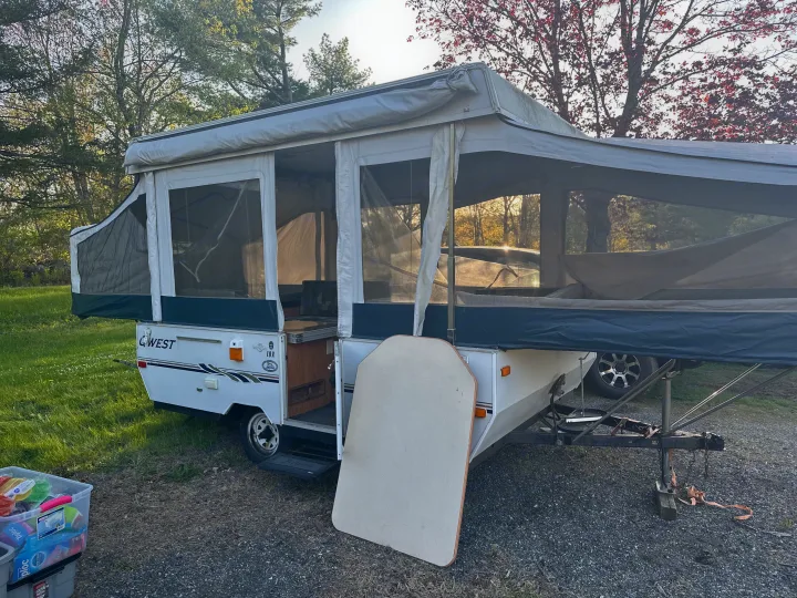 An open pop-up camper, an RV rental, fully set up in a wooded campsite with green grass and gravel.. Jayco Qwest 2004