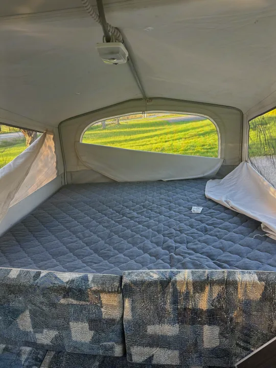 Camping RV rental pop-up interior: comfy gray bed, patterned seating, bright window view of green grass.. Jayco Qwest 2004