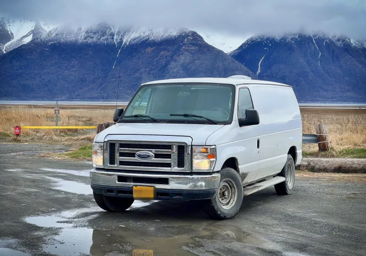 White Ford cargo van, a great rv rental option, parked on a wet road with snowy mountains in background.. Ford Econolline 250s 2013