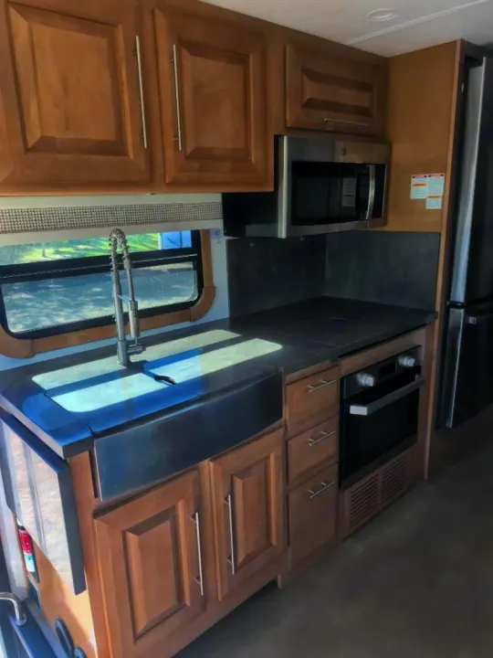 Modern RV rental kitchen featuring light wood cabinets, a large stainless steel sink, microwave, and built-in oven.. Forest River GEORGETOWN 36D7 2024