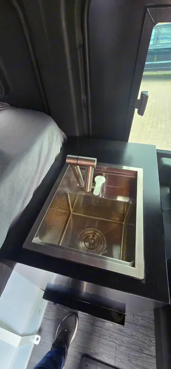 Inside an RV rental: sleek stainless steel sink with faucet next to a cozy bed. Dark interior with a window view.. Mercedes Sprinter 2023