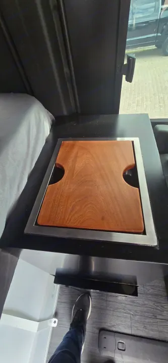 This RV rental's compact kitchen features a light wood cutting board integrated into a black countertop, with a bed nearby.. Mercedes Sprinter 2023