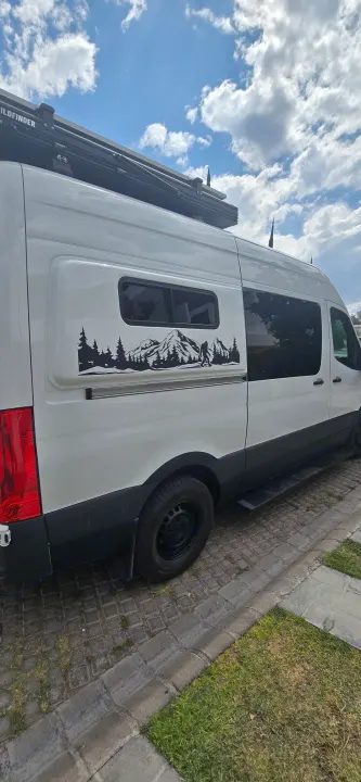 Adventure-ready white rv rental van with a custom Bigfoot mountain decal and roof rack, parked curbside under blue sky.. Mercedes Sprinter 2023