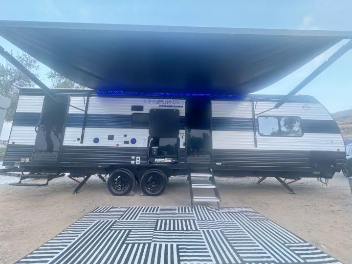 Cherokee Grey Wolf rv rental with extended awning, blue LEDs, outdoor kitchen, steps, and striped mat on gravel.. Forest River 26KF 2026