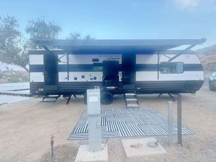 This RV rental is parked lakeside with an extended awning, blue lights, and a black/white striped mat.. Forest River 26KF 2026
