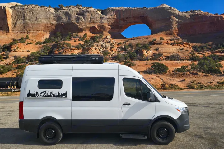 White Sprinter RV rental enjoying epic red rock scenery & natural arch. Bigfoot decal adds adventure to this camper van.. Mercedes Sprinter 2023