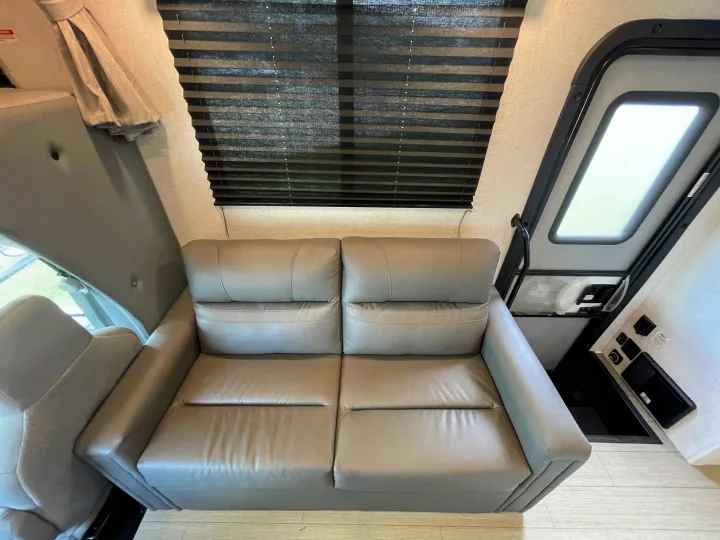 Inside an RV rental, showing a grey two-seater sofa, window with dark blinds, and an entry door with controls.. Entrada Entrada Motorhome 2022