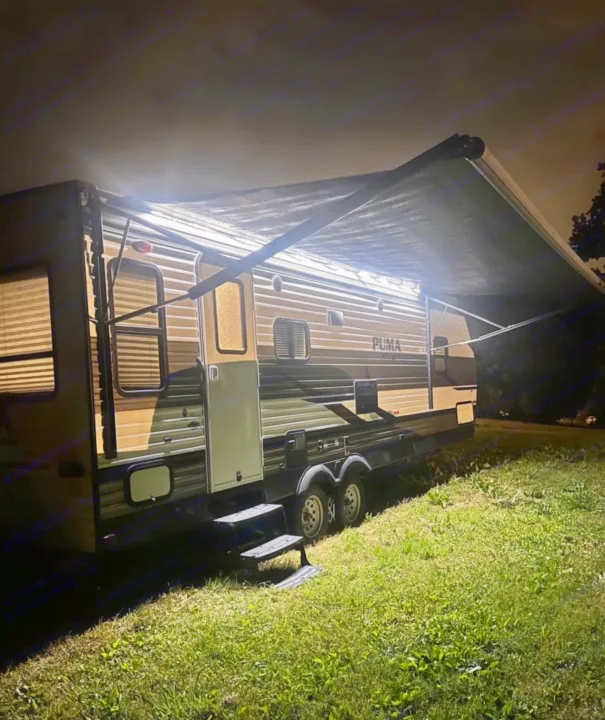Cozy night scene: a well-lit RV rental, a travel trailer with an extended awning, parked on grass. Perfect for an evening camp.. Forest River Palomino Puma XLE 2023