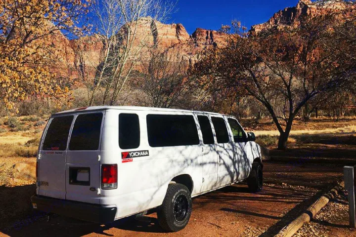 White custom van parked on a dirt path, surrounded by autumn trees and red rock mountains. Ideal for an RV rental adventure.. Ford E350 2000