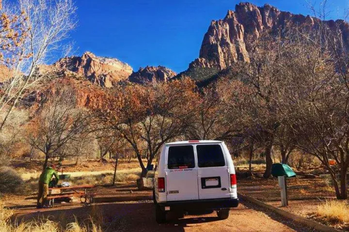 White campervan (RV rental) at a stunning desert campsite with majestic red rock mountains and bare trees. Perfect for adventure!. Ford E350 2000