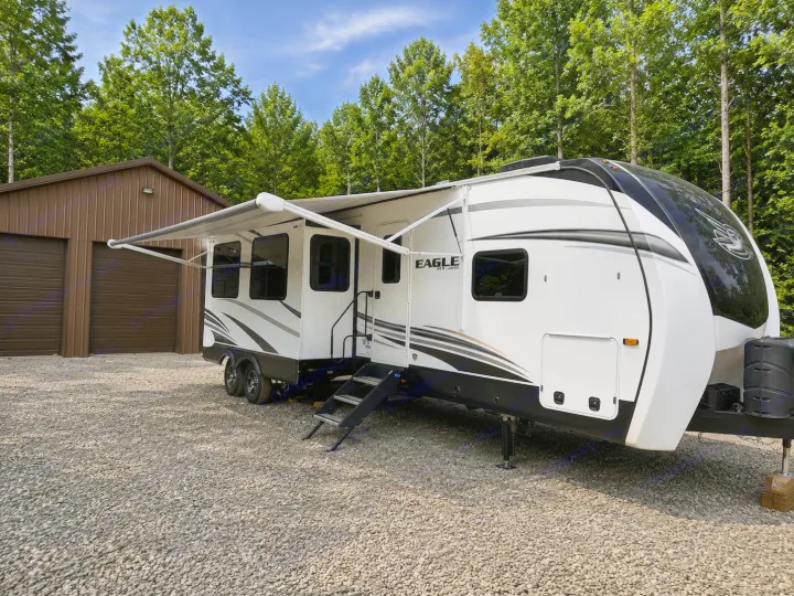White & black Eagle by Jayco fifth wheel RV rental on a gravel lot. Awning out, steps down, beside a brown garage and trees.. Jayco Eagle HT 2022