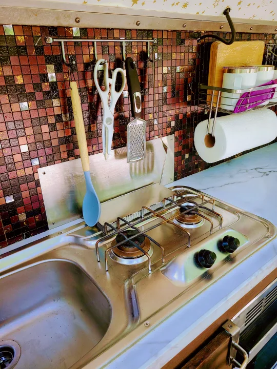 Compact RV rental kitchen with a stainless steel sink, two-burner stove, tiled backsplash, and hanging utensils.. nuCamp T@G 2019