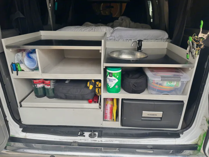 Organized RV rental kitchen module with sink, Dometic fridge, propane, and cleaning supplies. Bed visible in background.. Ford Econoline 150 2012