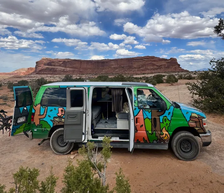 Vibrantly painted RV rental camper van parked with doors open amidst red rock canyons under a cloudy sky.. Ford Econoline 150 2012