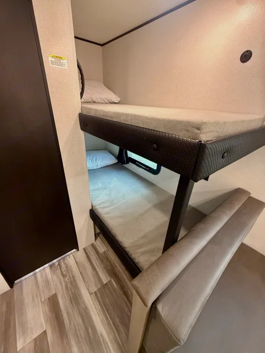 An RV rental interior features compact bunk beds with light gray mattresses, a dark brown cabinet, and plush bench seating on wood-look floors.. Jayco SLX 175BH 2026