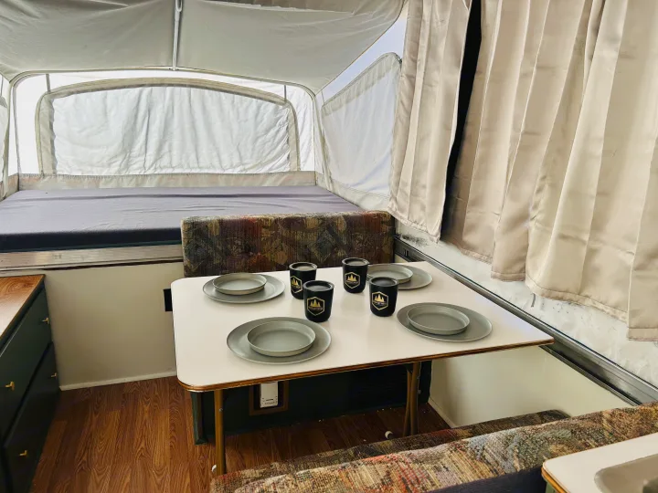 Interior of an RV rental pop-up camper with a set dining table, patterned bench seating, and a bed area.. Coleman Destiny Santa Fe 1996