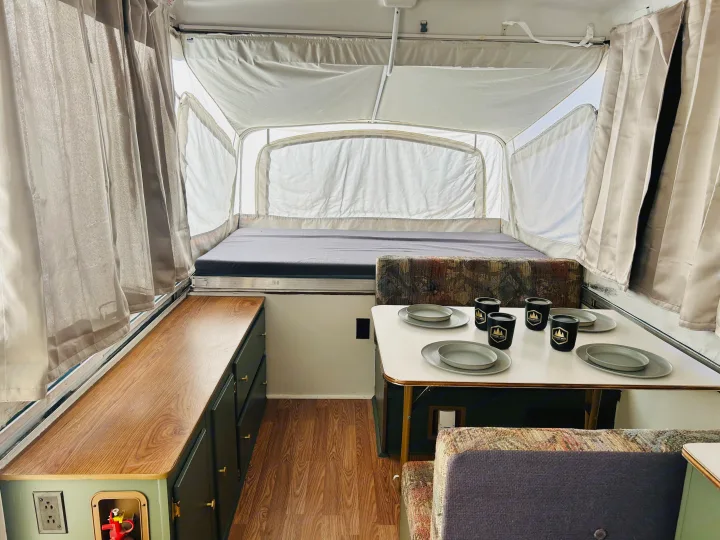 Inside a bright pop-up rv rental, featuring a bed, dining table with dishes, and cabinet storage.. Coleman Destiny Santa Fe 1996