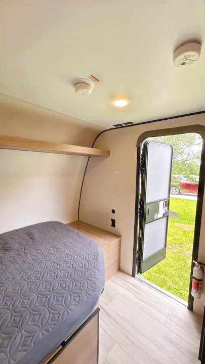 RV rental interior: cozy bed, wooden shelf, smoke alarms, fire extinguisher by the open door to the outdoors.. Keystone RV 13B travel Trailer 2026