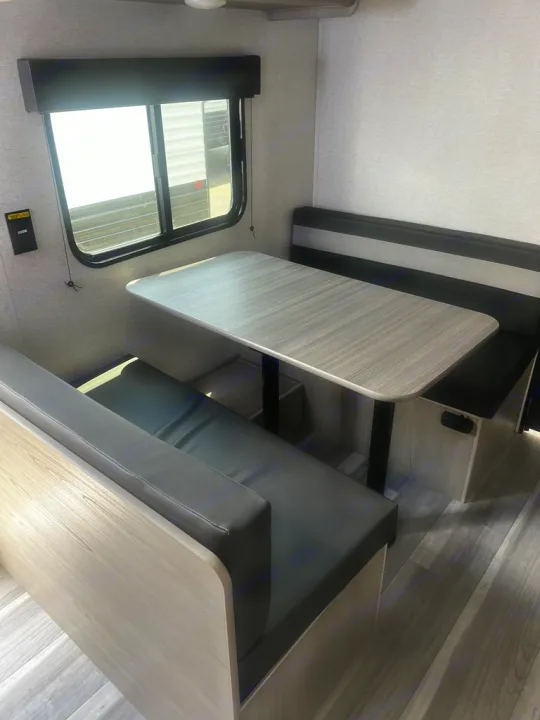 Modern RV rental dinette with light wood table & dark bench seating, by a window. Ideal for comfortable travel trailer living.. Keystone RV Springdale 2024