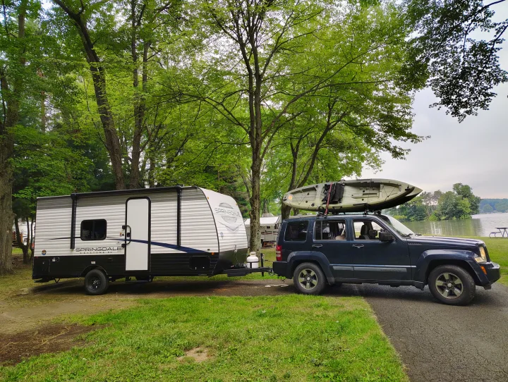 An RV rental with a kayak-laden SUV at a scenic lakeside campsite under lush trees, ready for outdoor adventures.. Keystone RV Springdale 2024