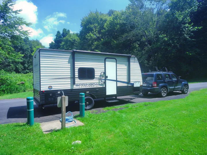 This Springdale travel trailer RV rental, towed by a Jeep SUV, is fully setup at a sunny campsite for outdoor camping.. Keystone RV Springdale 2024