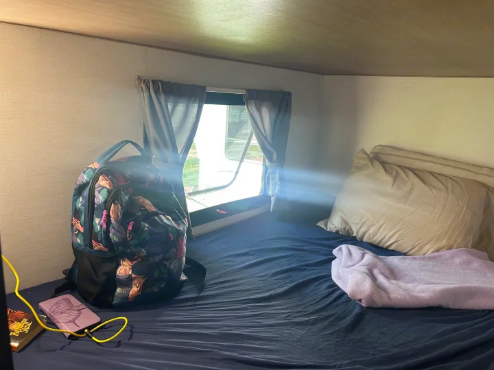 An RV rental sleeping area shows a bed, patterned backpack, book, and a vivid ray of sunlight streaming from the window.. Gulf Stream Grand River 25BH 2026