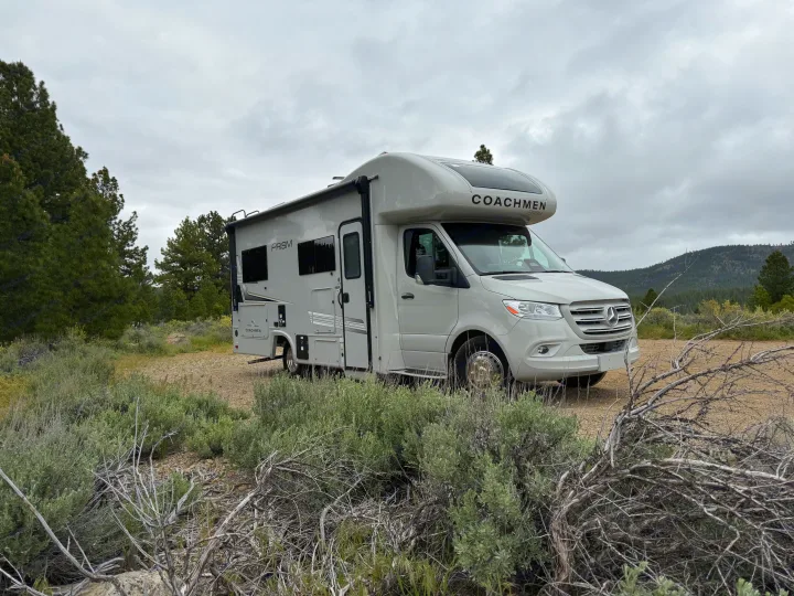 This light grey Coachmen Prism RV rental is parked among sagebrush and pine trees, ready for adventure under a cloudy sky.. Coachmen Prism 2026