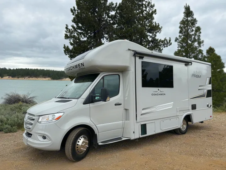 Scenic rv rental: a grey Coachmen Prism motorhome parked on a dirt path next to a calm lake with pine trees under an overcast sky.. Coachmen Prism 2026