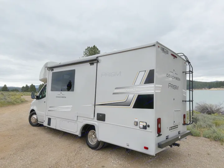 Class C Coachmen Prism RV rental on a dirt path by a lake, under cloudy skies. Great for camping and outdoor travel.. Coachmen Prism 2026