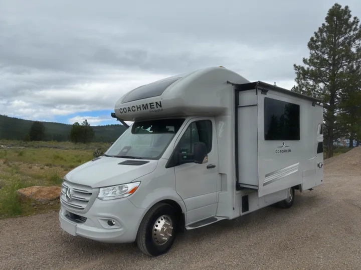 Light grey Coachmen Prism Class C RV rental, parked on gravel. Forested hills and a cloudy sky provide a scenic backdrop.. Coachmen Prism 2026