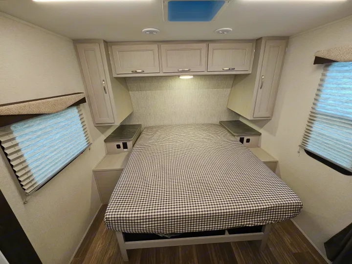 Cozy RV rental bedroom with a checkered comforter on the bed, overhead cabinets, side tables, and windows.. Cruiser RV Shadow Cruiser 2025