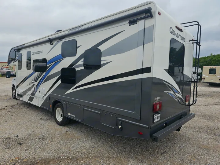 A white, grey, and blue Quantum RV rental is parked on a gravel lot, featuring an awning and a rear ladder.. Thor Quantum 2023