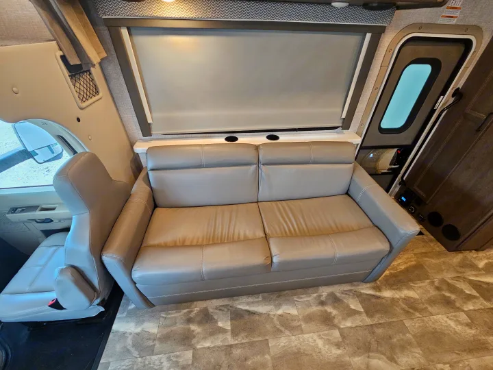 RV rental living space: modern sofa with cupholders, window blind, and driver's seat. Ready for adventure.. Thor Quantum 2023