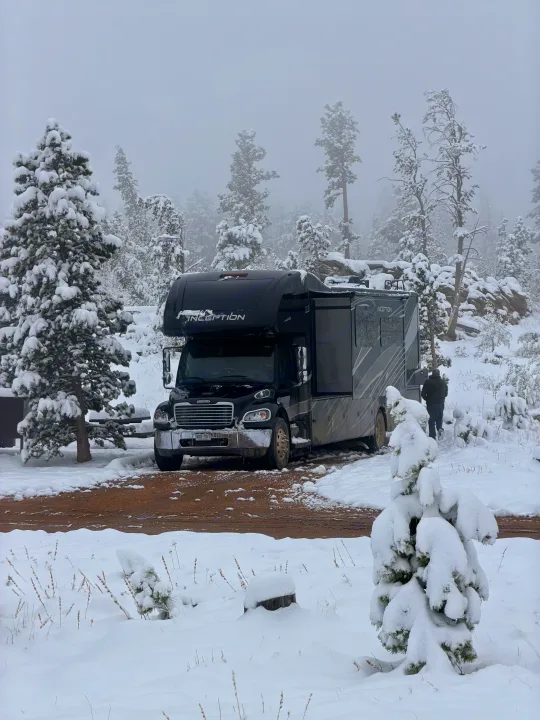 A black RV rental is parked among snow-covered pine trees in a winter forest campsite. A person walks in the snowy background.. Thor Inception 2023