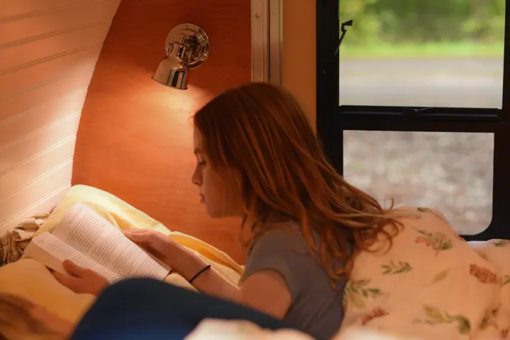 Cozy RV rental interior: A young person reads a book in bed by a warm light, with a window view to nature.. All Good Equipment Company Teardrop Trailer 2026