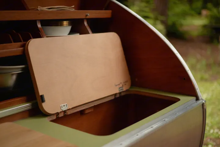 Interior of an RV rental camper: wooden kitchen with dishes, a hidden storage compartment, and olive green accents, ready for adventure.. All Good Equipment Company Teardrop Trailer 2026