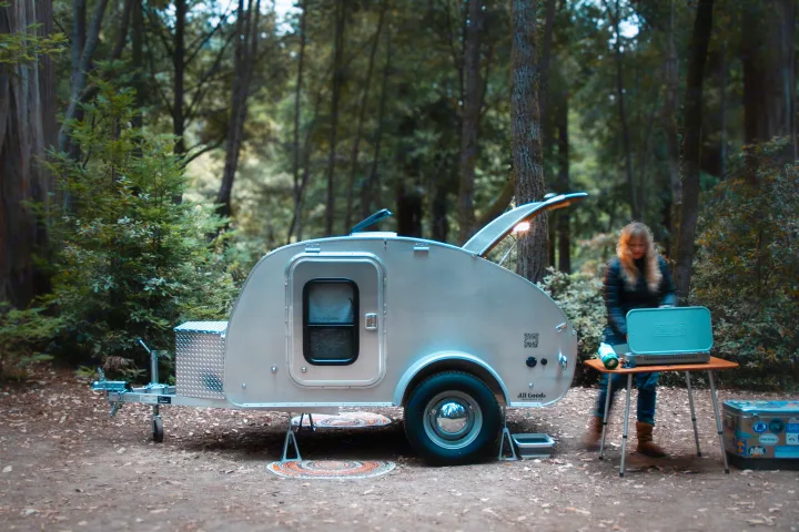 Camping setup with a sleek teardrop RV rental parked among tall trees. A person prepares a meal on a stove next to it.. All Good Equipment Company Teardrop Trailer 2026