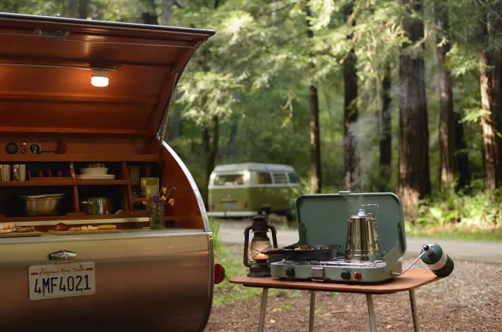 An RV rental teardrop trailer kitchen with camping stove, boiling kettle, and a vintage VW van parked in a serene forest.. All Good Equipment Company Teardrop Trailer 2026