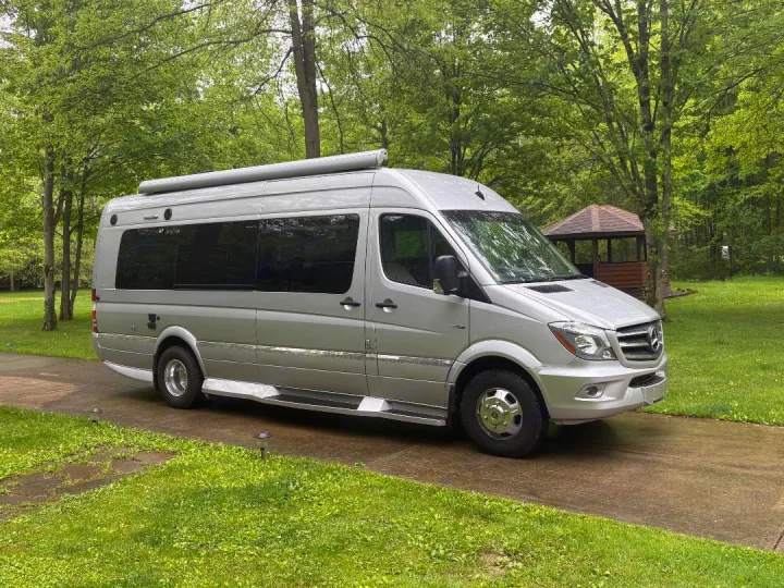 This silver Mercedes Sprinter RV rental, a sleek camper van, is parked on a wet paved path surrounded by lush trees.. Winnebago Era 2017