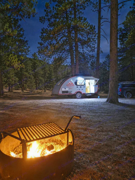 Cozy rv rental at a starry night campsite with a warm campfire in the foreground, surrounded by tall pine trees.. Liberty Outdoors Little Guy 2018