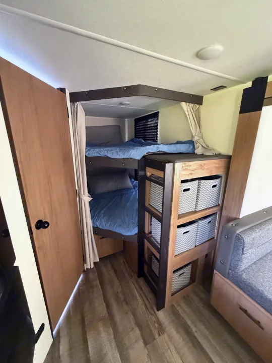 RV rental interior featuring bunk beds with blue bedding, a storage unit with patterned baskets, and wood-look flooring.. Forest River Campsite Reserve 2025