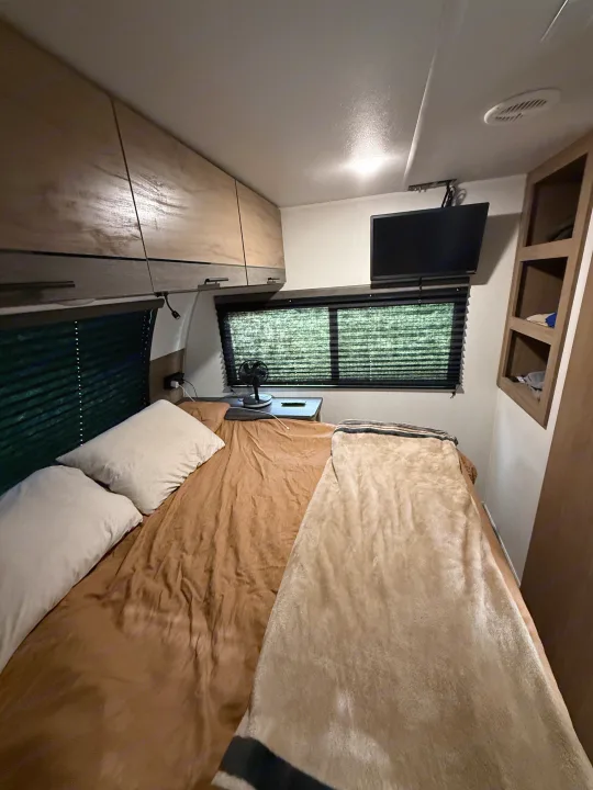 Cozy RV rental bedroom featuring a bed with pillows and blankets, overhead cabinets, a window with blinds, and a mounted TV.. Forest River Campsite Reserve 2025
