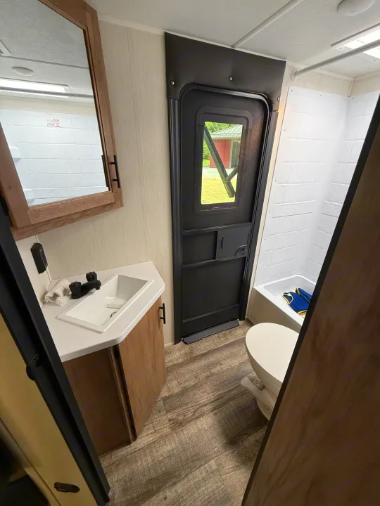 Compact RV rental bathroom: a sink, vanity, toilet, and small shower with a blue item inside. Mirror above sink, wood-look floor.. Forest River Campsite Reserve 2025