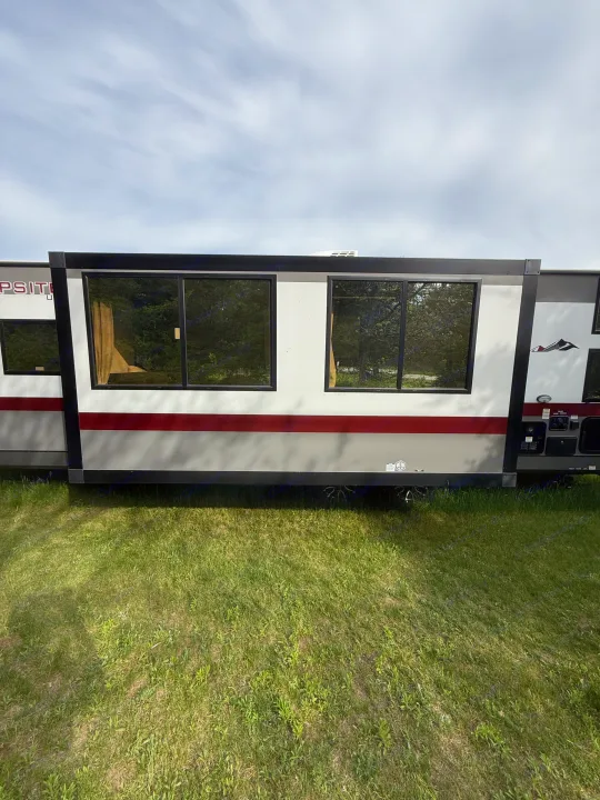 RV rental side view: A white, grey & red travel trailer with large windows, parked on green grass under a cloudy sky.. Forest River Campsite Reserve 2025
