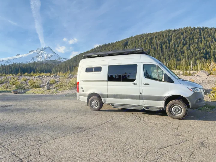 A silver rv rental camper van parked on cracked asphalt, facing a majestic snow-capped mountain and green forest.. Mercedes-Benz Sprinter 2020
