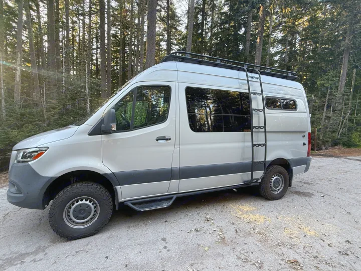 Silver Mercedes Sprinter camper van with off-road tires & roof rack in a forest. Ideal RV rental for outdoor adventures.. Mercedes-Benz Sprinter 2020