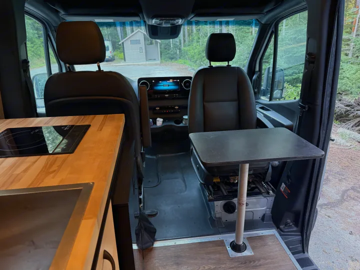rv rental camper van interior: kitchenette, swivel seat with table, and outdoor view of an outhouse & trees.. Mercedes-Benz Sprinter 2020