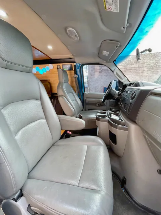 Interior of an RV rental van, showing gray front seats, dashboard, and custom-built wood paneling in the rear.. Ford E350 2010
