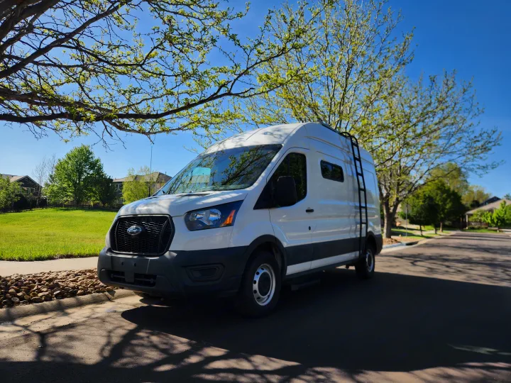 White Ford Transit high-roof RV rental van with a side ladder, parked on a sunny street under leafy trees and a blue sky.. Ford Transit 2022